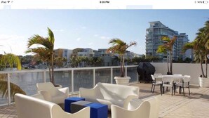 A rooftop terrace with white sofas, a blue ottoman, and a table with chairs. There is a view of a body of water and buildings in the background. - Spectacular Views, Great Location on Millionaire's Row (Miami Beach)
