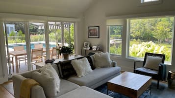 Un salon spacieux avec un canapé gris, une table basse et un fauteuil. On y trouve également une table à manger avec des chaises et une plante en pot. De grandes fenêtres offrent une vue sur une piscine et sur la verdure.