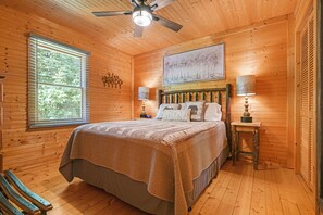 Ein Schlafzimmer einer Holzhütte mit einem Bett, einem Deckenventilator, einem Fenster mit Jalousien und einem Bild an der Wand.