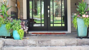 A house entrance with a "WELCOME" sign above the door, a stone step leading up to the door, and two large potted plants on either side.