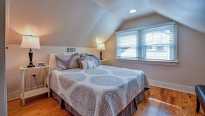 Quarto com cama grande, duas mesas de cabeceira com luminárias, janela com persianas e piso de madeira.