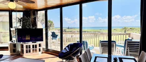 A spacious balcony with a wooden ceiling, large windows offering a view of the beach, a television, and outdoor seating.