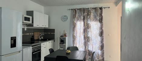 A compact kitchen with white cabinets, a microwave, and a water dispenser. There is a dining table with chairs and a clock on the wall.