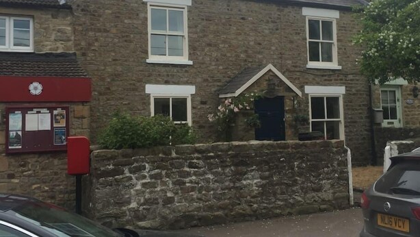 A stone building with a traditional design, featuring a central entrance with a dark door and white-framed windows. - Beautiful Stone Cottage in Wensleydale (Hunton, Bedale)