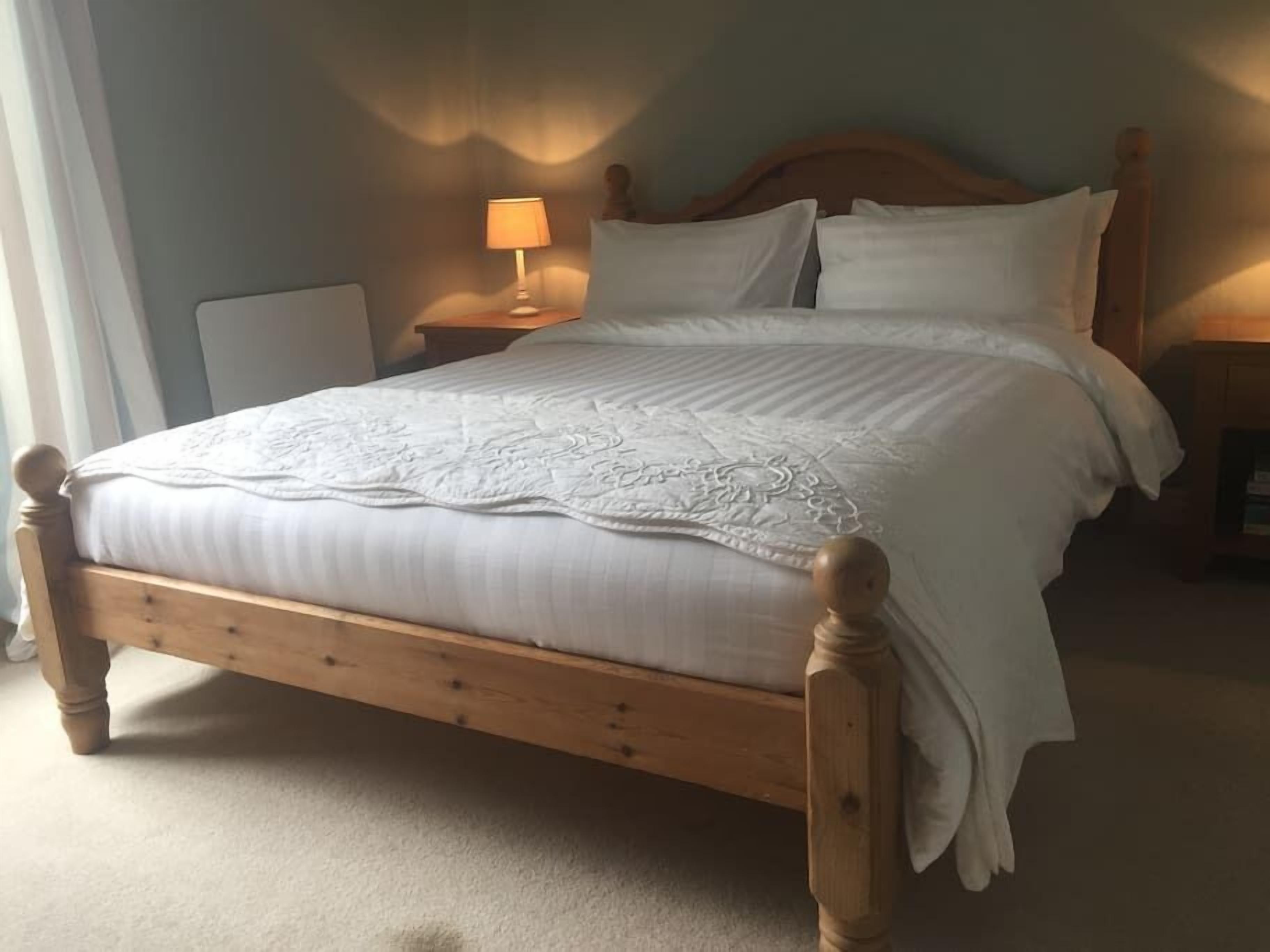 Un lit bien fait, avec une structure en bois, une literie blanche et une couverture décorative. Une lampe de chevet est posée sur une table de chevet en bois.