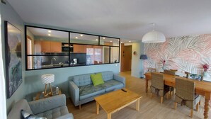 Ein modernes Wohnzimmer mit Sofa, Couchtisch und Essbereich mit Stühlen. Durch ein großes Fenster ist eine Küche zu sehen.