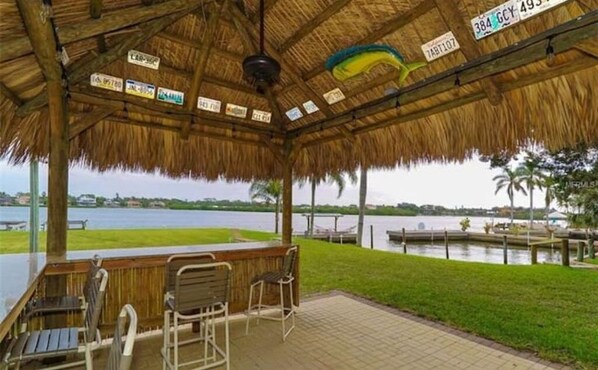 A covered outdoor area with a wooden table and chairs, a thatched roof, and a view of a body of water.