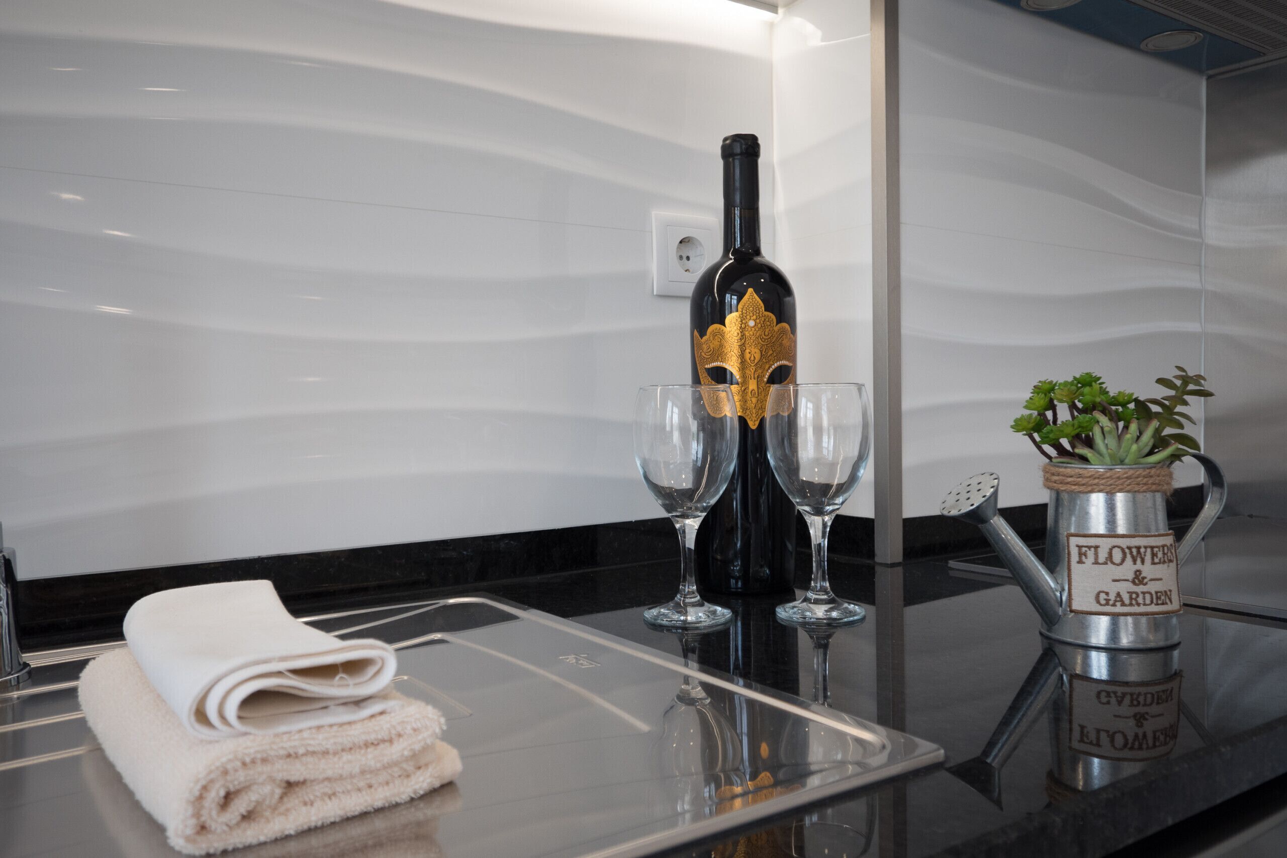 Une salle de bain avec une serviette, une bouteille de vin, deux verres et une plante dans un arrosoir en métal.