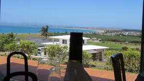 A table with a bottle and glasses, overlooking a coastal view with a house and greenery. - Stunning Views Over Tokerau Beach (Karikari Peninsula)