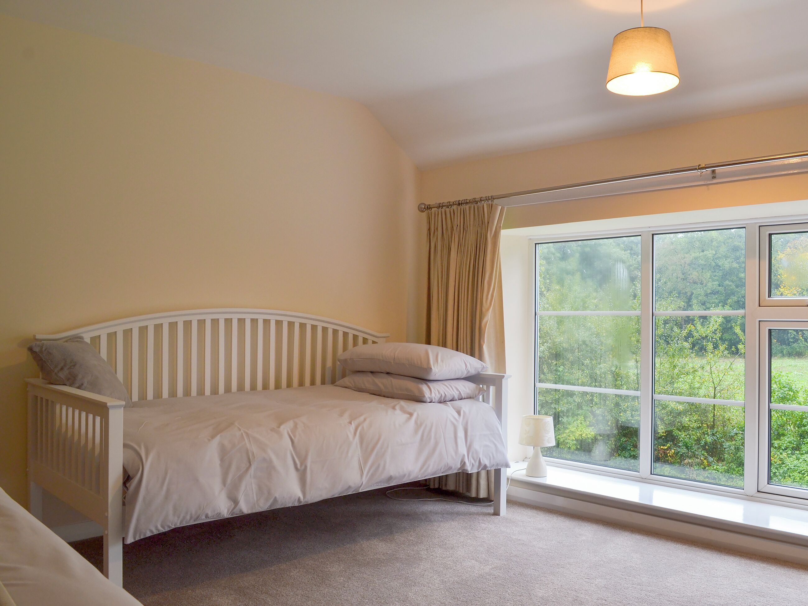 A bedroom with a single bed, a window with a view of greenery, and a hanging light fixture.