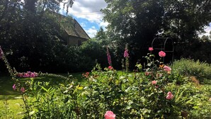Ein Garten mit verschiedenen Pflanzen und Blumen, darunter Rosen und hohe Blüten, mit einem Haus im Hintergrund.