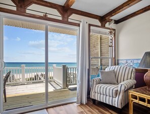Una habitació amb una gran porta corredissa de vidre que dona a un balcó de fusta amb vista a una platja.