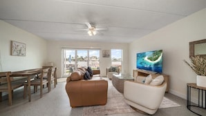 A spacious living room with a large TV, a brown leather sofa, a white sofa, and a dining area with wooden furniture.