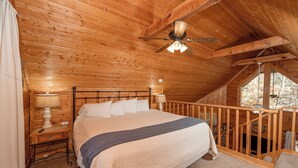 A wooden cabin bedroom with a large bed, two bedside lamps, a ceiling fan, and a window with a view of trees. - Cute, Cozy Cabin for a Romantic Getaway! Pool, Putt Putt, Fishing, Golf, Tennis (Sevierville)