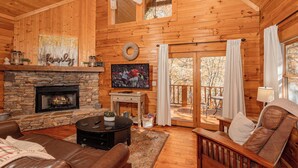 A cozy cabin interior with a stone fireplace, leather sofas, a television, and a balcony with curtains. - Cute, Cozy Cabin for a Romantic Getaway! Pool, Putt Putt, Fishing, Golf, Tennis (Sevierville)