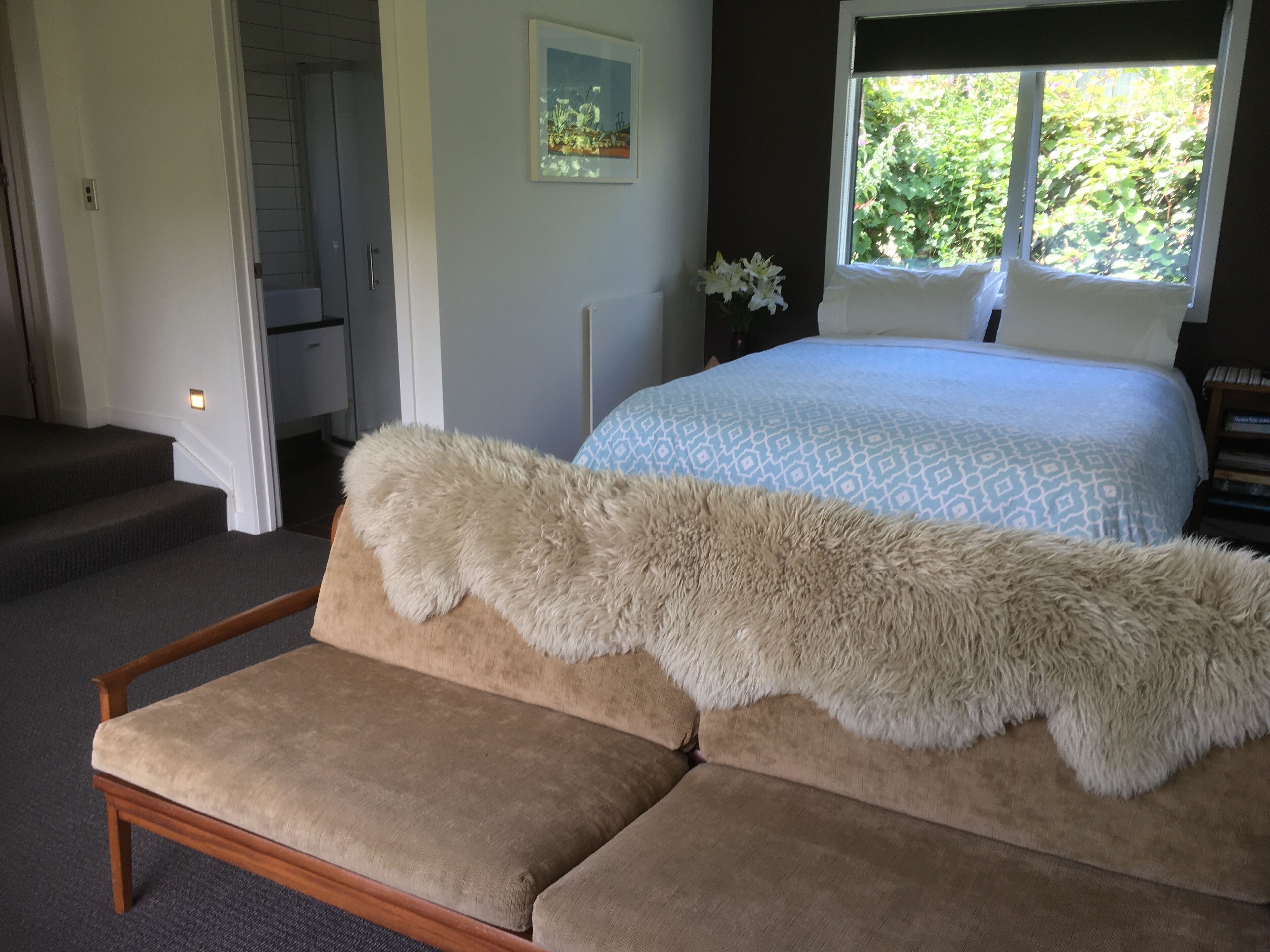 Een slaapkamer met een bed, een zachte witte plaid, een sofa, een schilderij en een raam met uitzicht op groen.