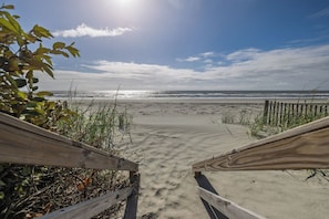 Un paseo marítimo de madera que conduce a una playa arenosa con cielo despejado y océano al fondo.