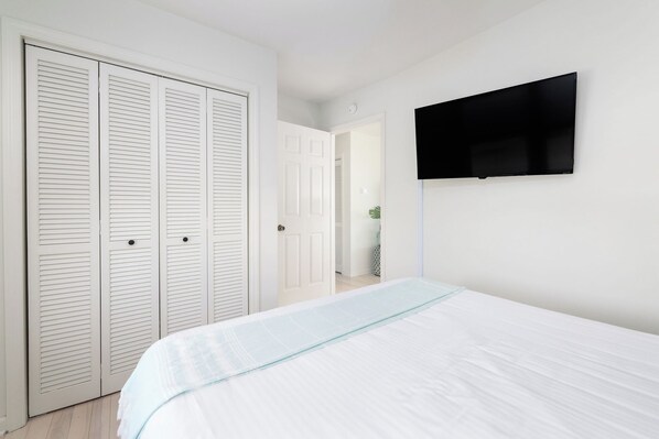 Un dormitorio con cama, televisión montada en la pared y un conjunto de armarios blancos con puertas a listones.