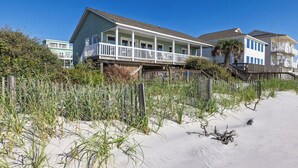 A beachfront house with a balcony, surrounded by sand and vegetation.