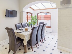 Un comedor con una mesa preparada para cuatro personas, rodeado de sillas de cuero, una planta en maceta y un televisor de pantalla plana montado en la pared.