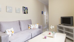A living room with a grey sofa, a flat-screen TV on a stand, a coffee table with a vase of flowers, and wall art.