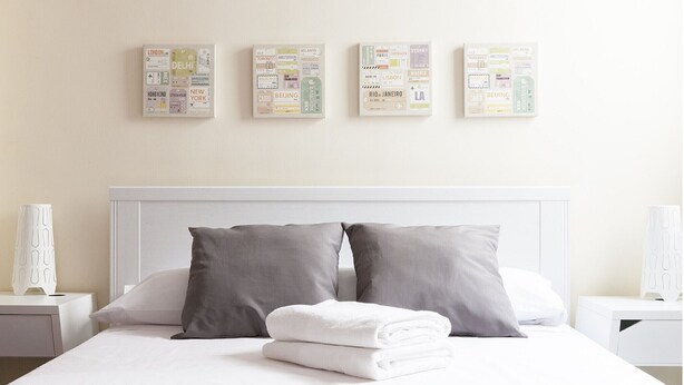A hotel room with a neatly made bed, grey pillows, and a white headboard. There are three framed pictures on the wall above the bed, each featuring various city names and landmarks.