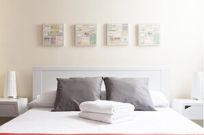 Una habitación de hotel con una cama bien hecha, almohadas grises y un cabecero blanco. Encima de la cama, en la pared, hay tres cuadros enmarcados, cada uno con diferentes nombres de ciudades y monumentos.