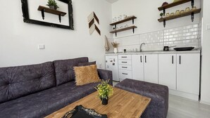 Ein modernes Wohnzimmer mit einem dunkelgrauen Sofa, einem Holztisch und einer Küchenzeile mit weißen Schränken und Holzregalen.