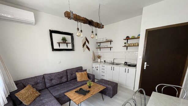 Ein modernes Wohnzimmer mit einer Eckcouch, einem Holztisch und einem Esstisch mit Stühlen. Es gibt eine Küchenzeile mit weißen Schränken und einer dunklen Tür.