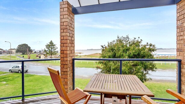 A balcony with a wooden table and chairs, a brick wall, and a view of a road and greenery.