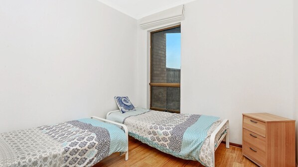 A small, bright room with two single beds, a wooden dresser, and a large window.