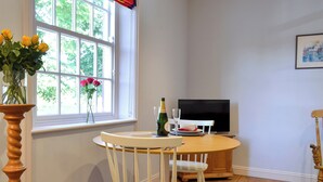 Ein Zimmer mit Holztisch, zwei weißen Stühlen, einem Fernseher und einem Fenster mit Blick auf Bäume.