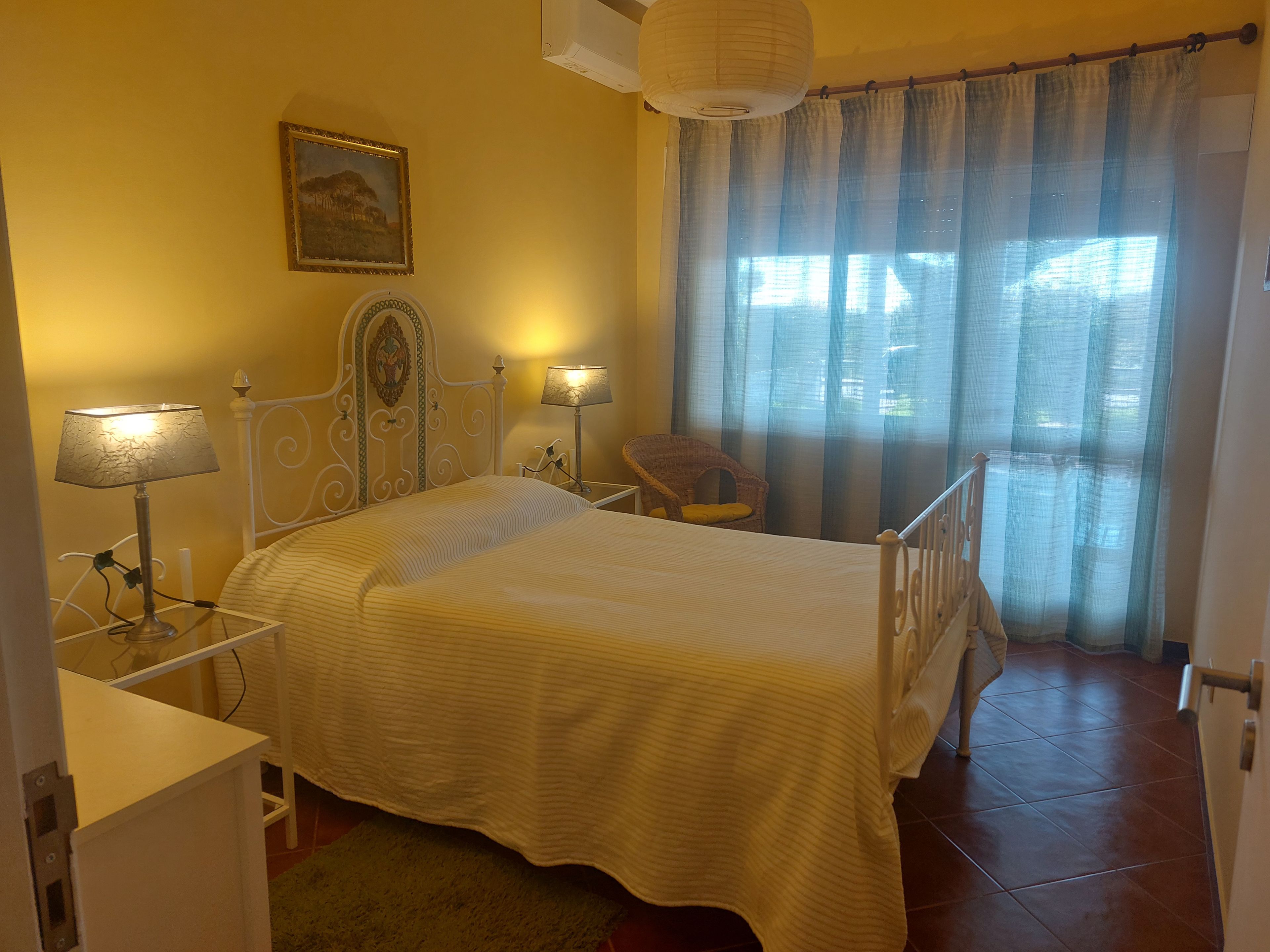 Un dormitorio con una cama, lámparas de noche, una silla, un cuadro y una ventana con cortinas.