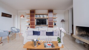 Un soggiorno moderno con un divano bianco, un tavolino da caffè con una bottiglia di champagne, bicchieri e snack, una libreria, una TV e un camino.
