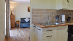 A compact kitchen with a sink, cabinets, and a countertop. There is a dining area with a sofa and a small table. - The Stables at Greenview (Winsham, near Chard)