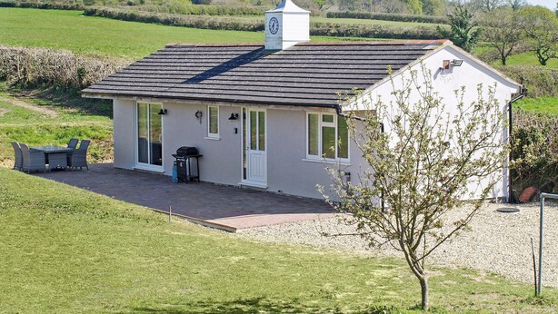 A small, single-story house with a sloped roof, a chimney, and a small porch with outdoor seating. - The Stables at Greenview (Winsham, near Chard)