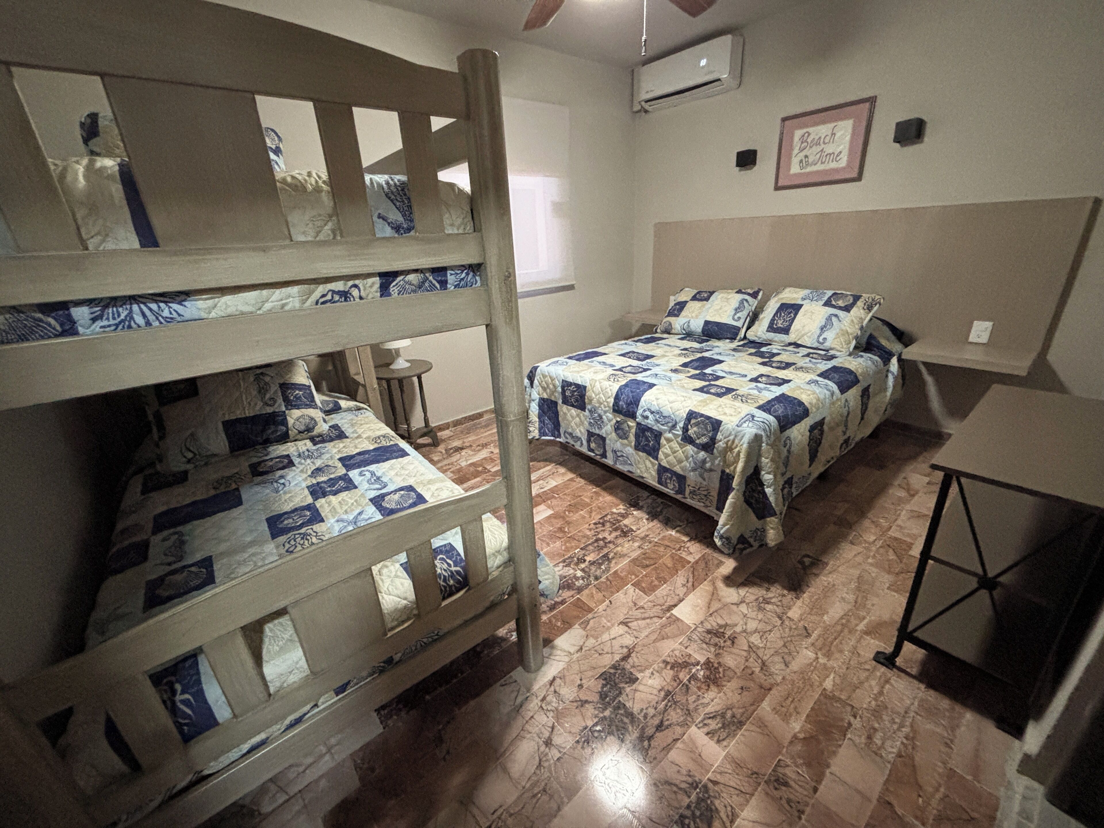 Een kamer met stapelbedden, een klein tafeltje, een stoel, een bed met quilt en een wandairconditioner, plus een lijst met een bordje.