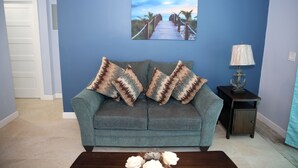 A living room with a blue wall, a grey sofa with patterned cushions, a wooden coffee table, and a framed picture of a beach walkway. - Cozy Eastern Beachfront Villas (Grand Bahama)