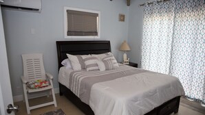 A bedroom with a bed, a chair, a nightstand, a lamp, a window, and a curtain. - Cozy Eastern Beachfront Villas (Grand Bahama)