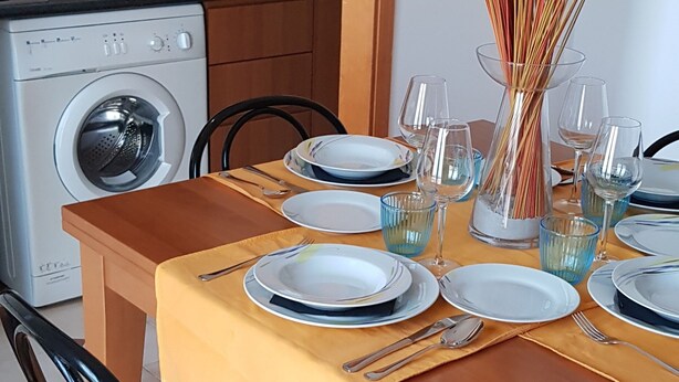 Een eettafel gedekt met borden, glazen en bestek, met een wasmachine en magnetron op de achtergrond.