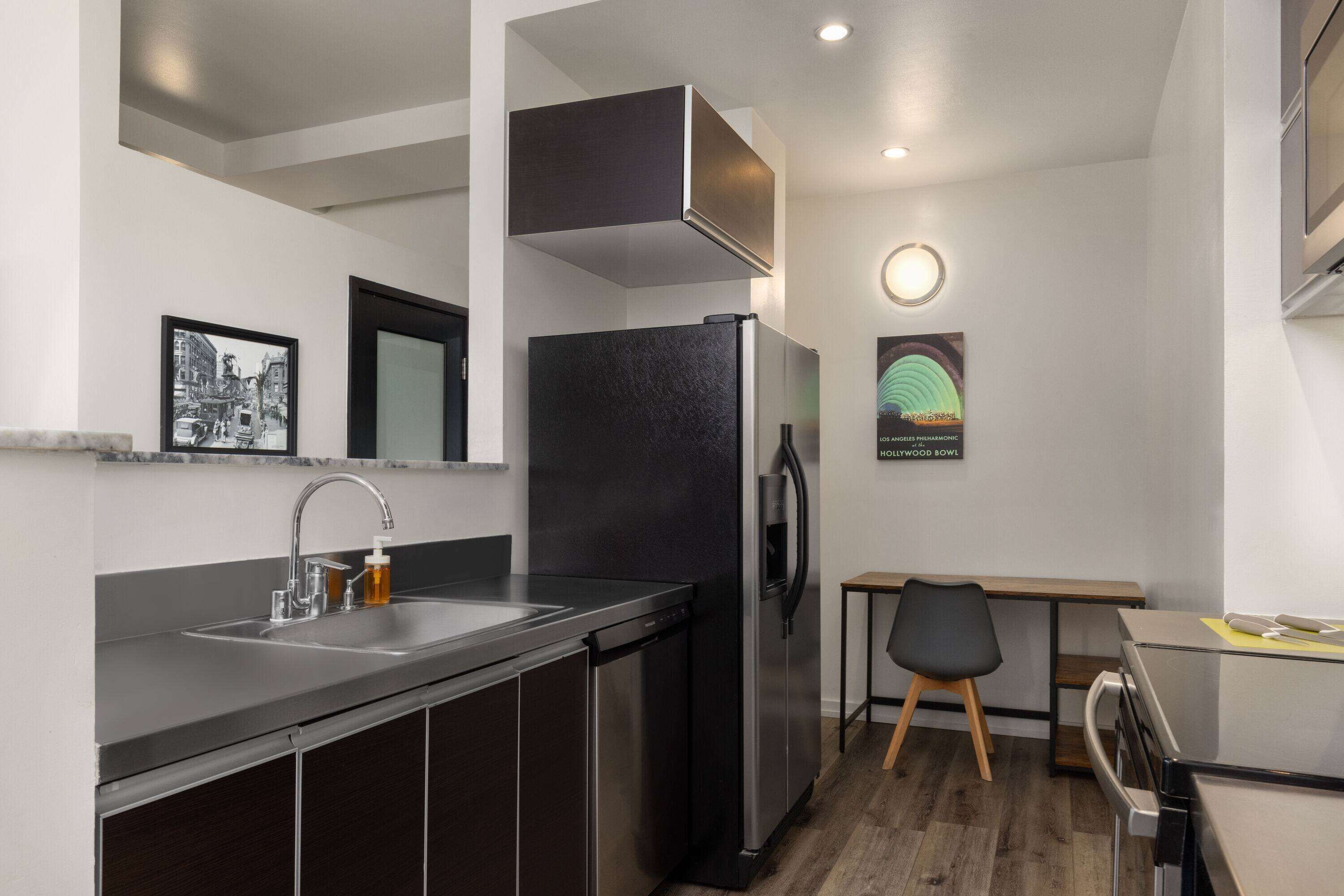 A compact kitchen with a stainless steel refrigerator, a sink, and a wooden dining table with a chair.