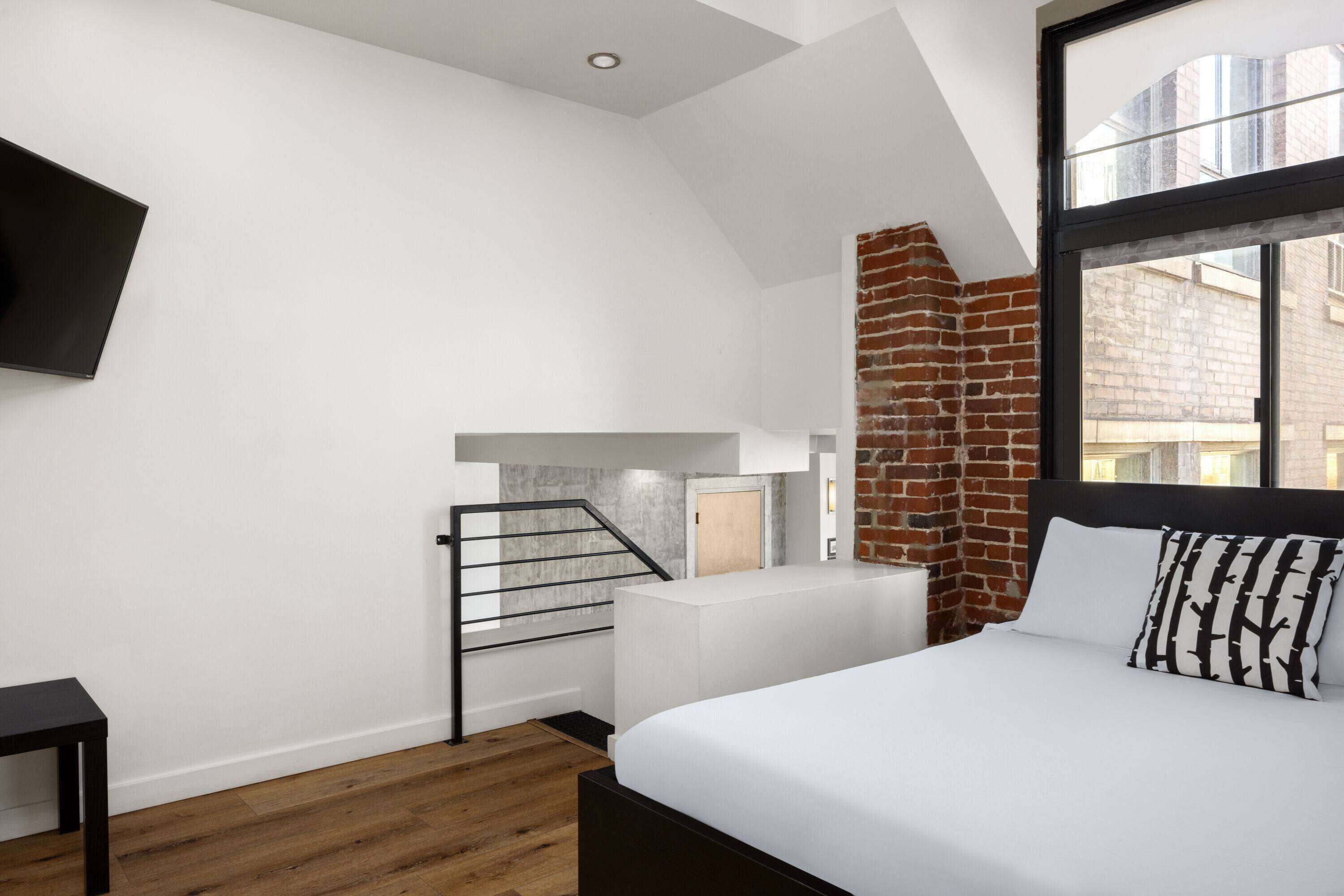 A modern bedroom with a large bed, a brick accent wall, a staircase, and a view of the city through a large window.