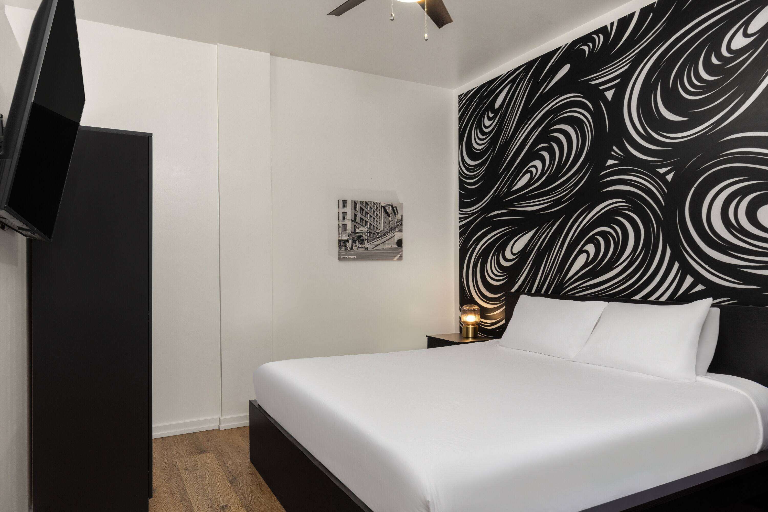 Une chambre d’hôtel moderne avec un grand lit, un téléviseur à écran plat fixé au mur, une petite table de chevet avec une lampe et une tête de lit à motifs noir et blanc.