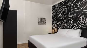 Une chambre d’hôtel moderne avec un grand lit, un téléviseur à écran plat fixé au mur, une petite table de chevet avec une lampe et une tête de lit à motifs noir et blanc.