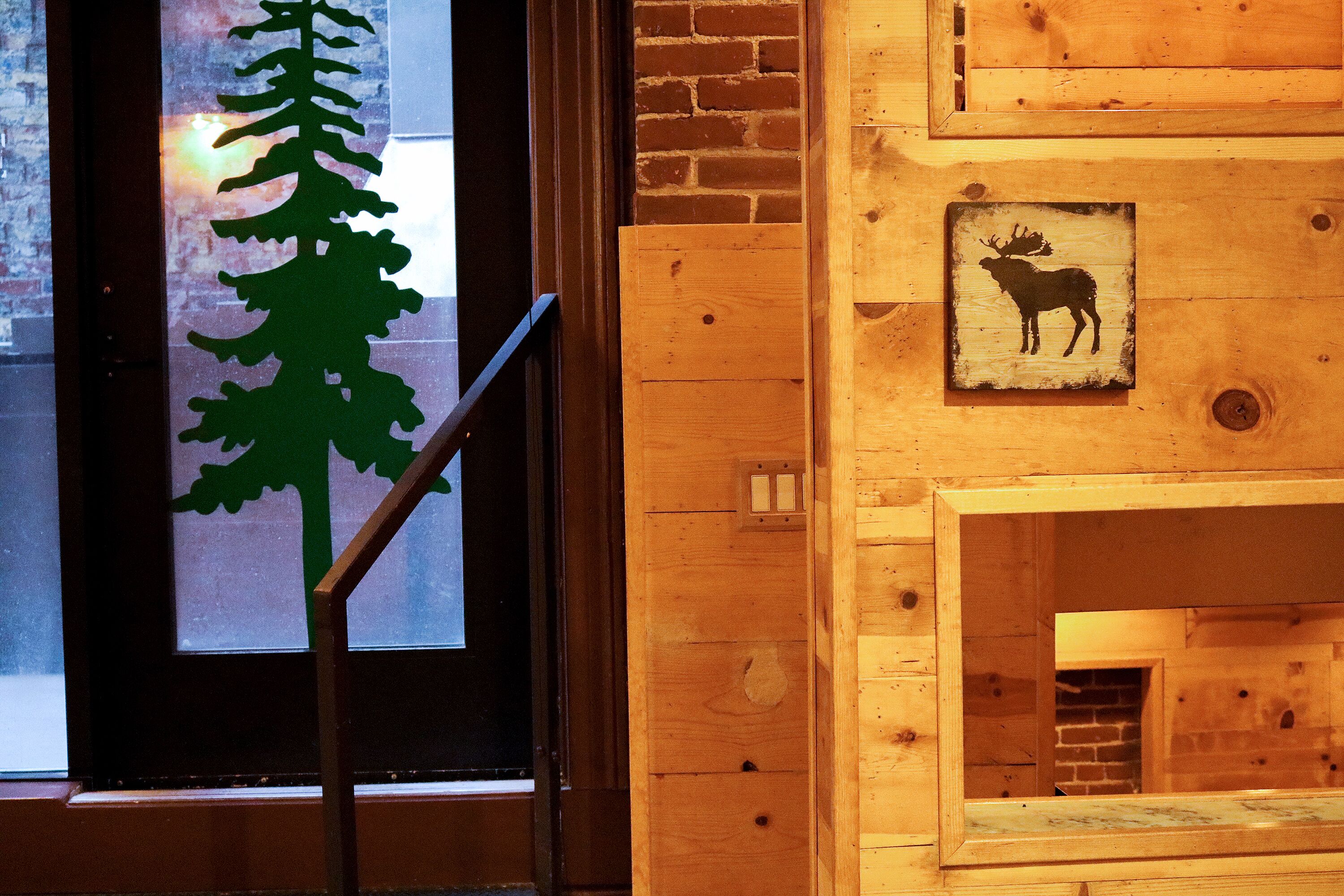 Interior de madera con una ventana que muestra la silueta de un árbol, una puerta de madera y un cartel con la imagen de un alce.