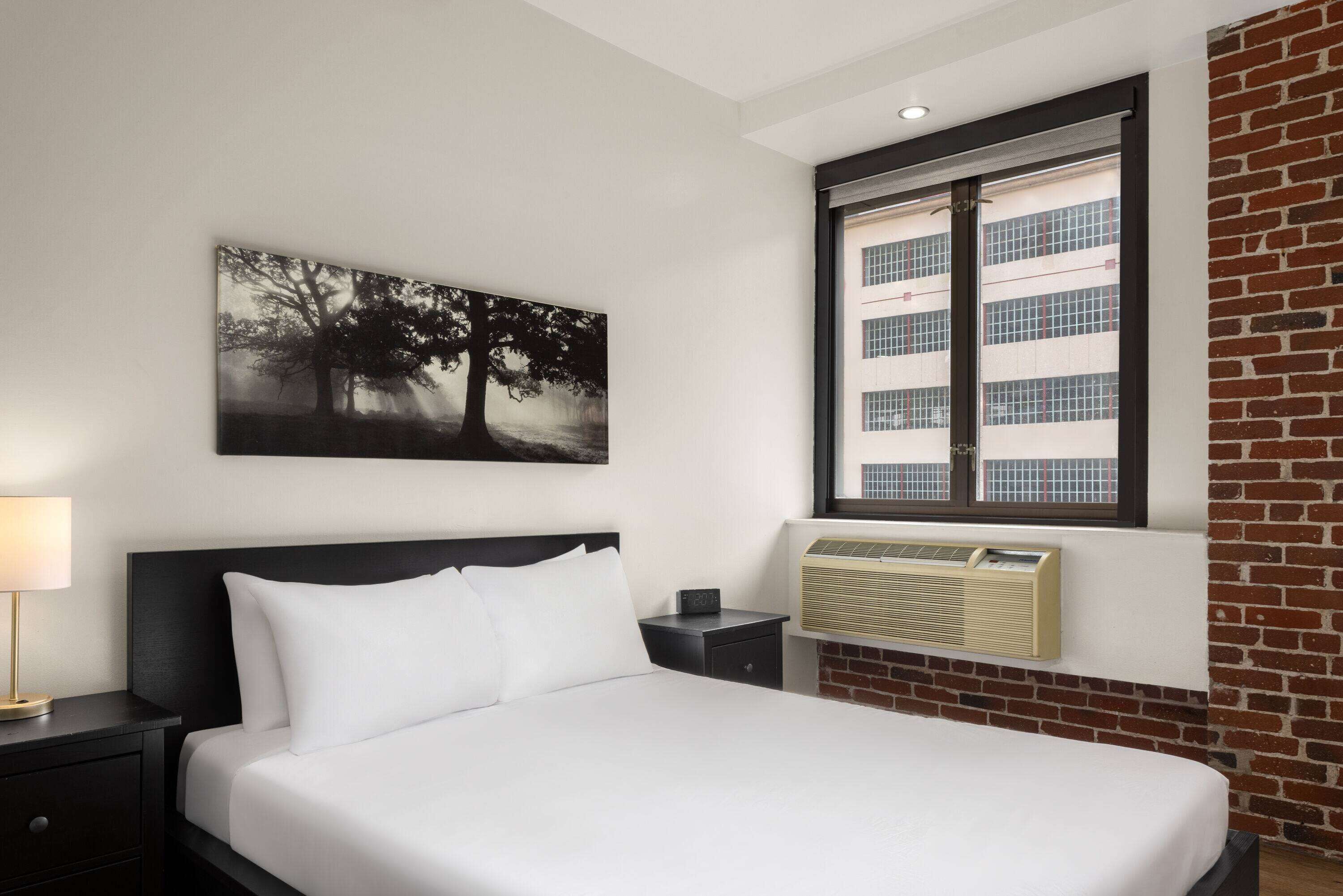 A hotel room with a bed, a nightstand, a lamp, a wall-mounted air conditioner, and a window with a view of a building.