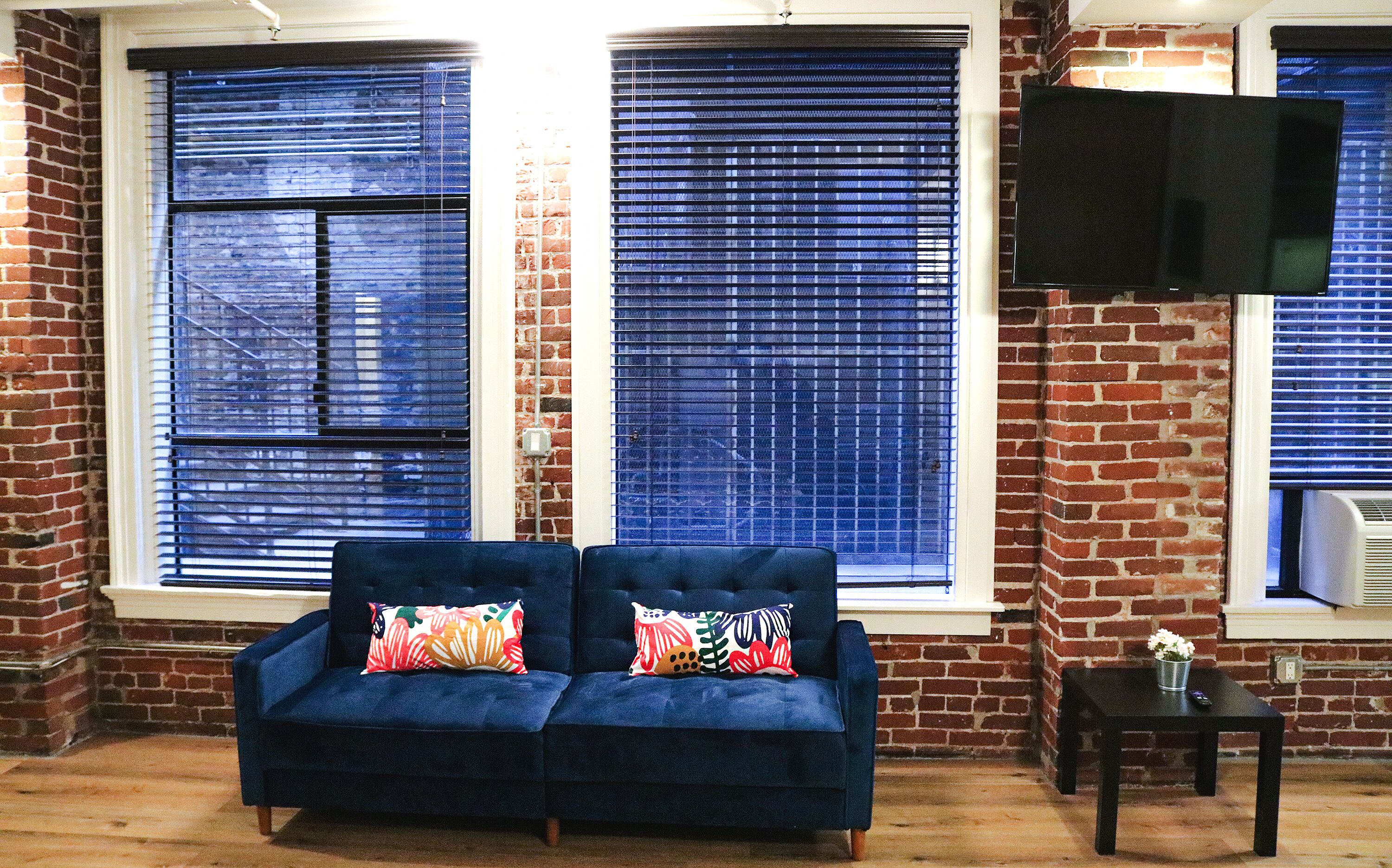 A living room with a dark blue sofa, two colorful pillows, and a TV on the wall.