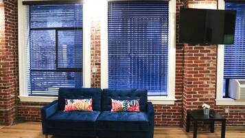 A living room with a dark blue sofa, two colorful pillows, and a TV on the wall.