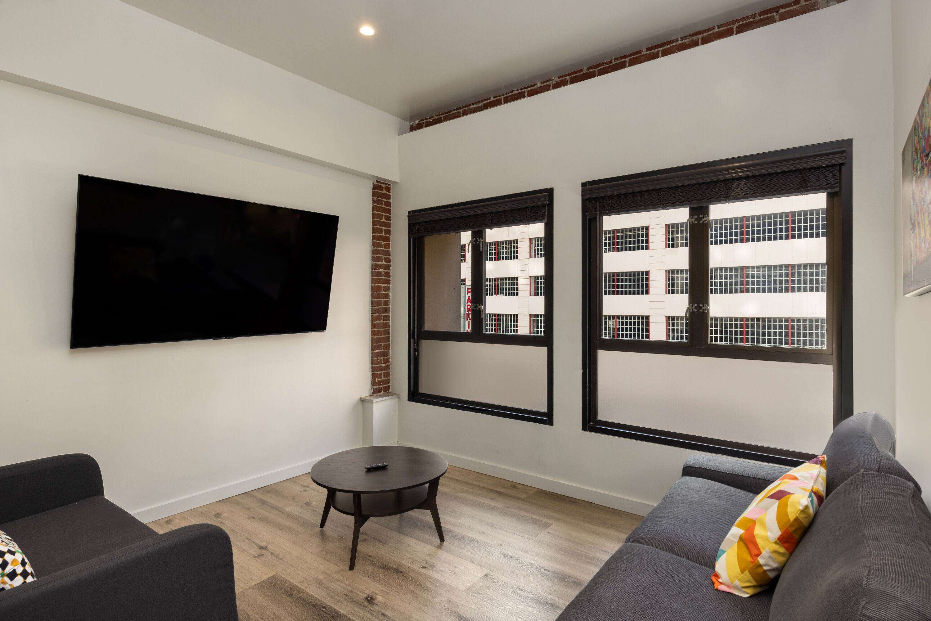 Una sala de estar moderna con un televisor de pantalla plana, una mesa de centro redonda y un sofá gris con un cojín colorido.