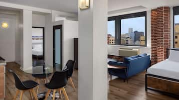 A modern apartment with a dining area, a living room with a sofa and a bed, and a large window with a city view.